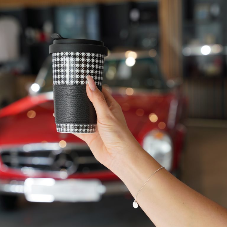 Zu sehen ist eine Hand die einen karierten Coffee to go Becher hält. Im Hintergrund sieht man verschwommen ein rotes Fahrzeug.
