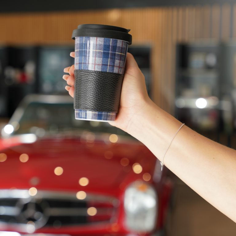 Zu sehen ist eine Hand die einen karierten Coffee to go Becher hält. Im Hintergrund sieht man verschwommen ein rotes Fahrzeug.