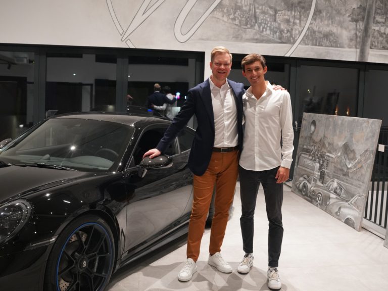 Zu sehen ist Rouven Genz mit einer weiteren Person vor einem schwarzen Fahrzeug im Ausstellungsraum von Wallner Classic