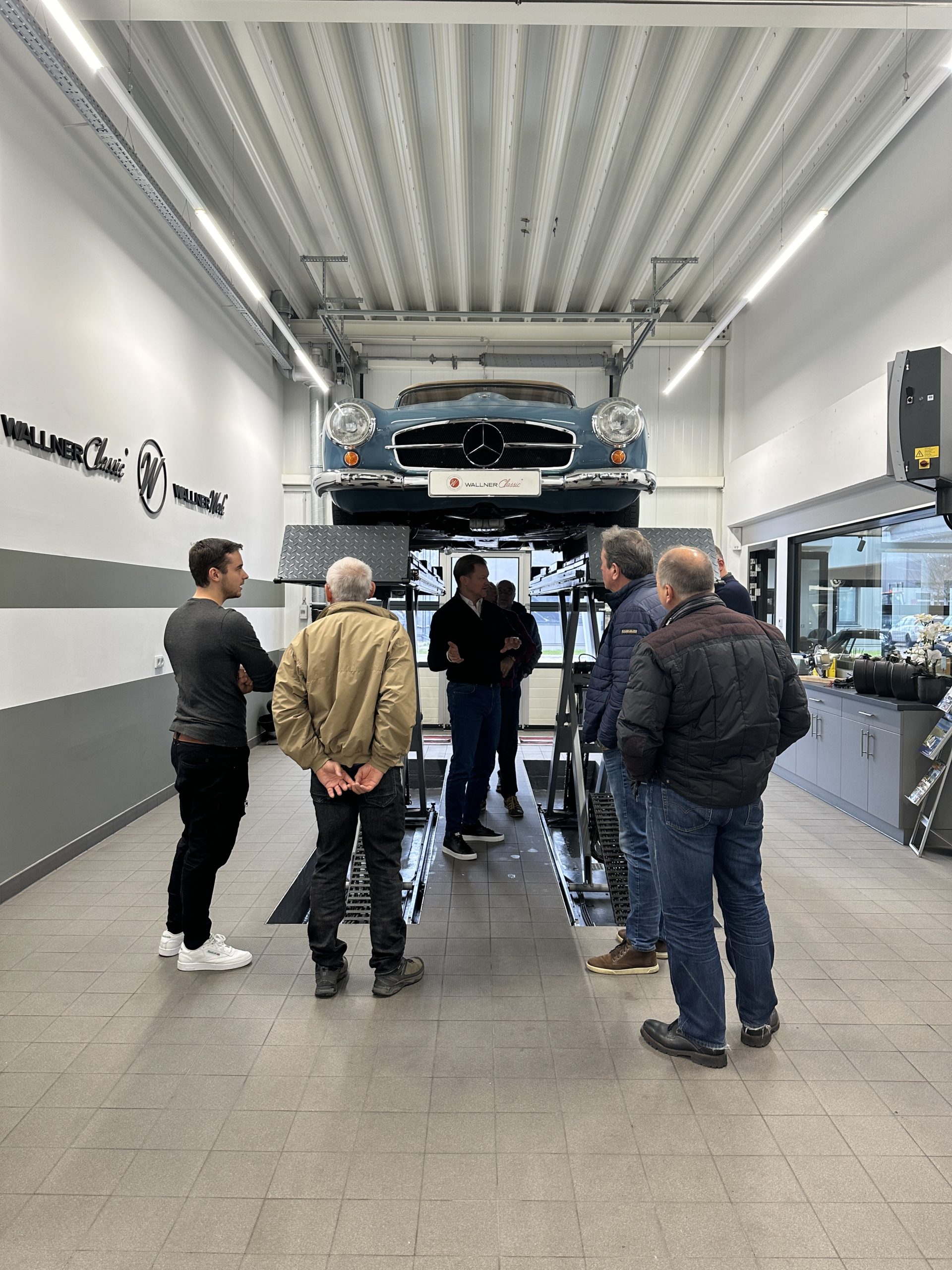 Zu sehen ist Herr Genz unter der Diagnosebühne. Ein blauer 190 SL befindet sich auf der Bühne. Weitere Personen begutachten das Fahrzeug.