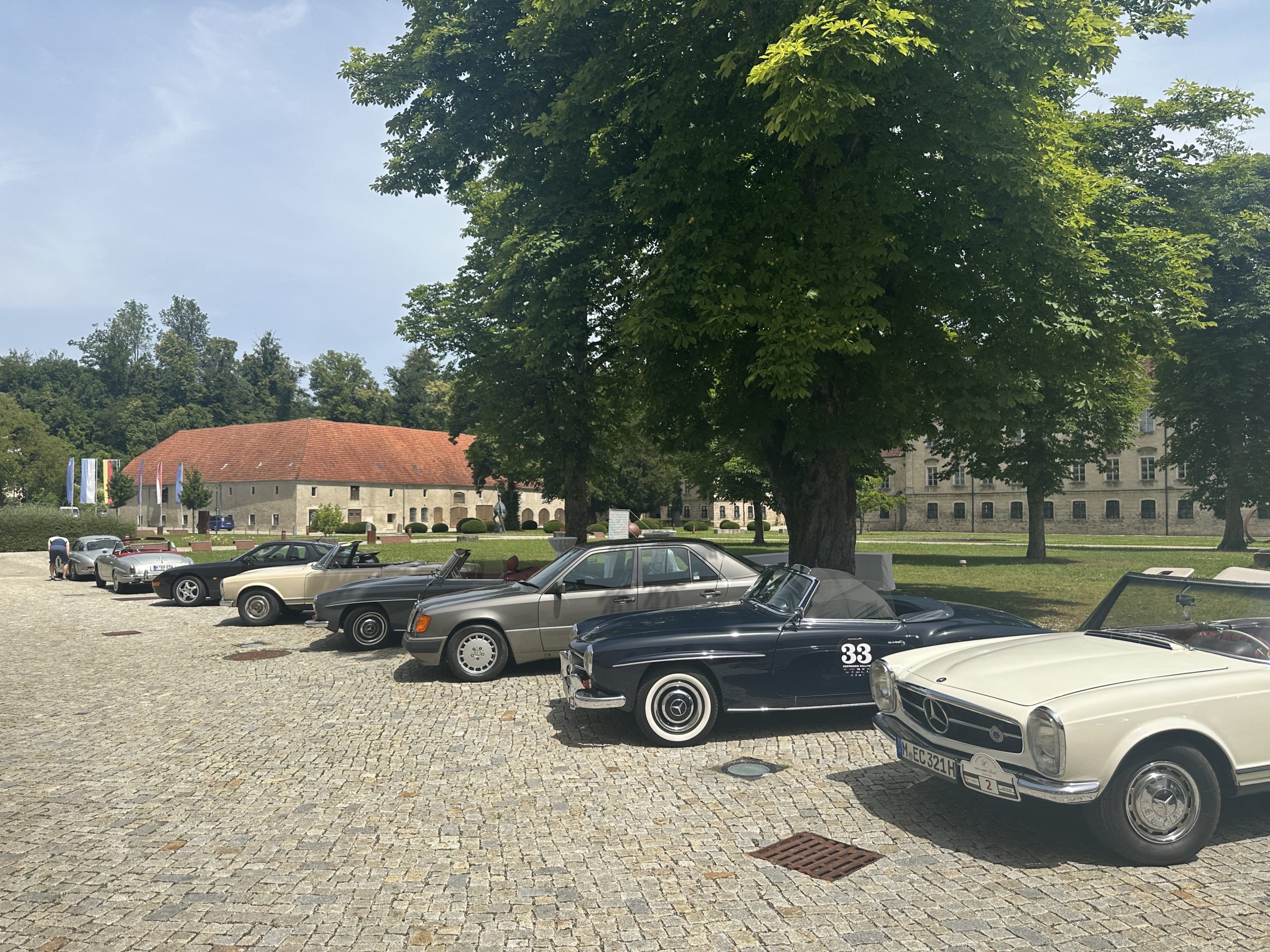 Zu sehen sind viele Oldtimer in dem Innenhof eines Guts. Des weiteren sieht man noch zwei große Kastanien Bäume