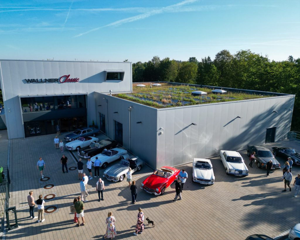 Auf dem Foto ist eine Luftaufnahme des Wallner Classic Werks. Vor dem Gebäude stehen mehrere Oldtimer Fahrzeuge, sowie Personen