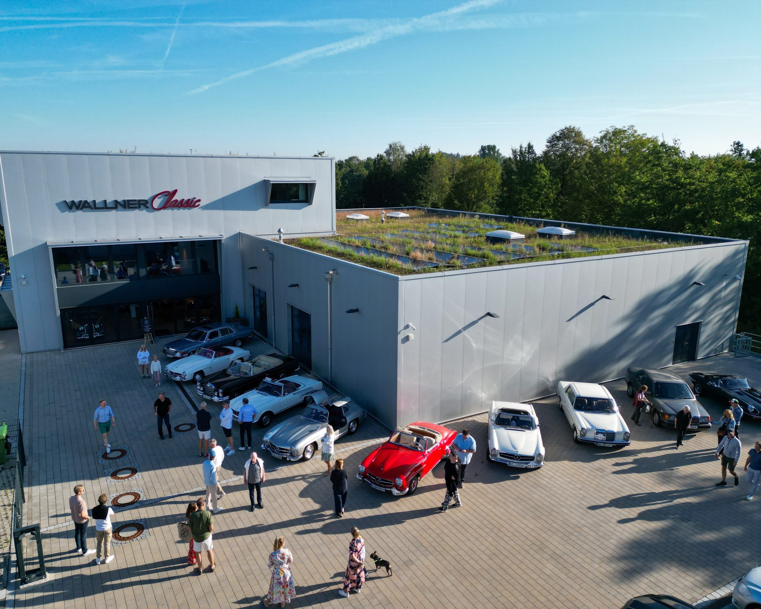 Auf dem Foto ist eine Luftaufnahme des Wallner Classic Werks. Vor dem Gebäude stehen mehrere Oldtimer Fahrzeuge, sowie Personen