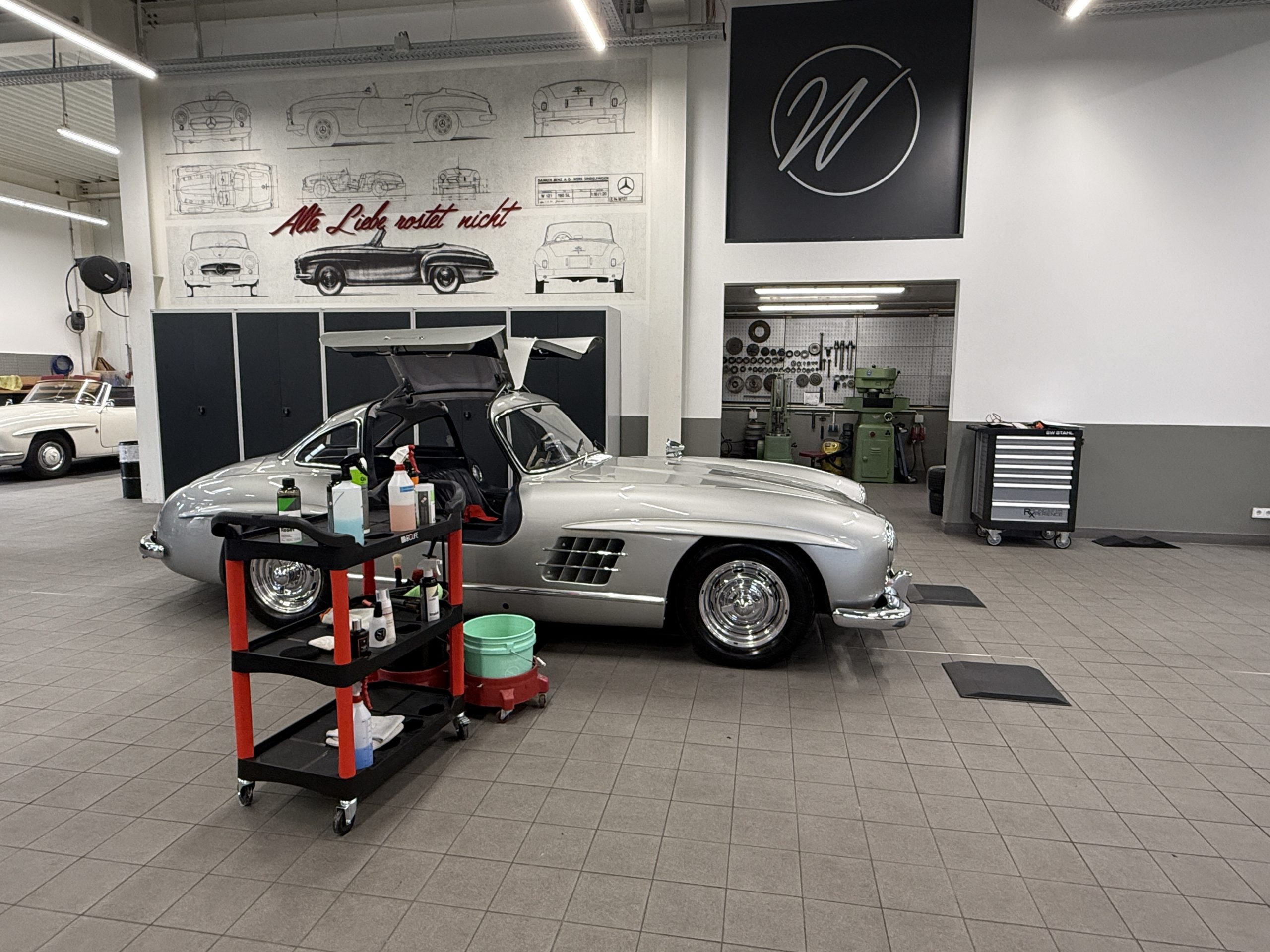 Zu sehen ist ein Mercedes 300 SL Flügeltürer, sowie ein Wagen mit Reinigungsmitteln. Das Foto wurde in der Wallner Classic Werkstatt aufgenommen
