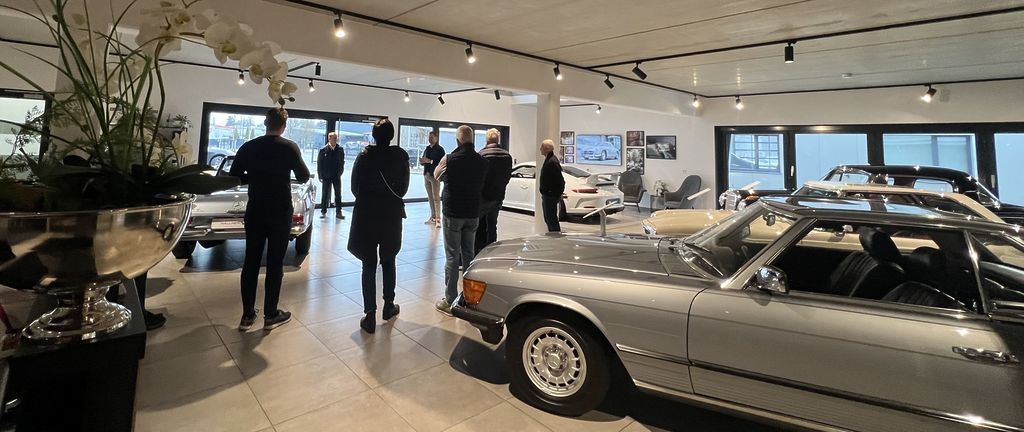Zu sehen ist ein Ausstellungsraum mit mehreren Mercedes Fahrzeugen und Kunden in einem Halbkreis aufgestellt.